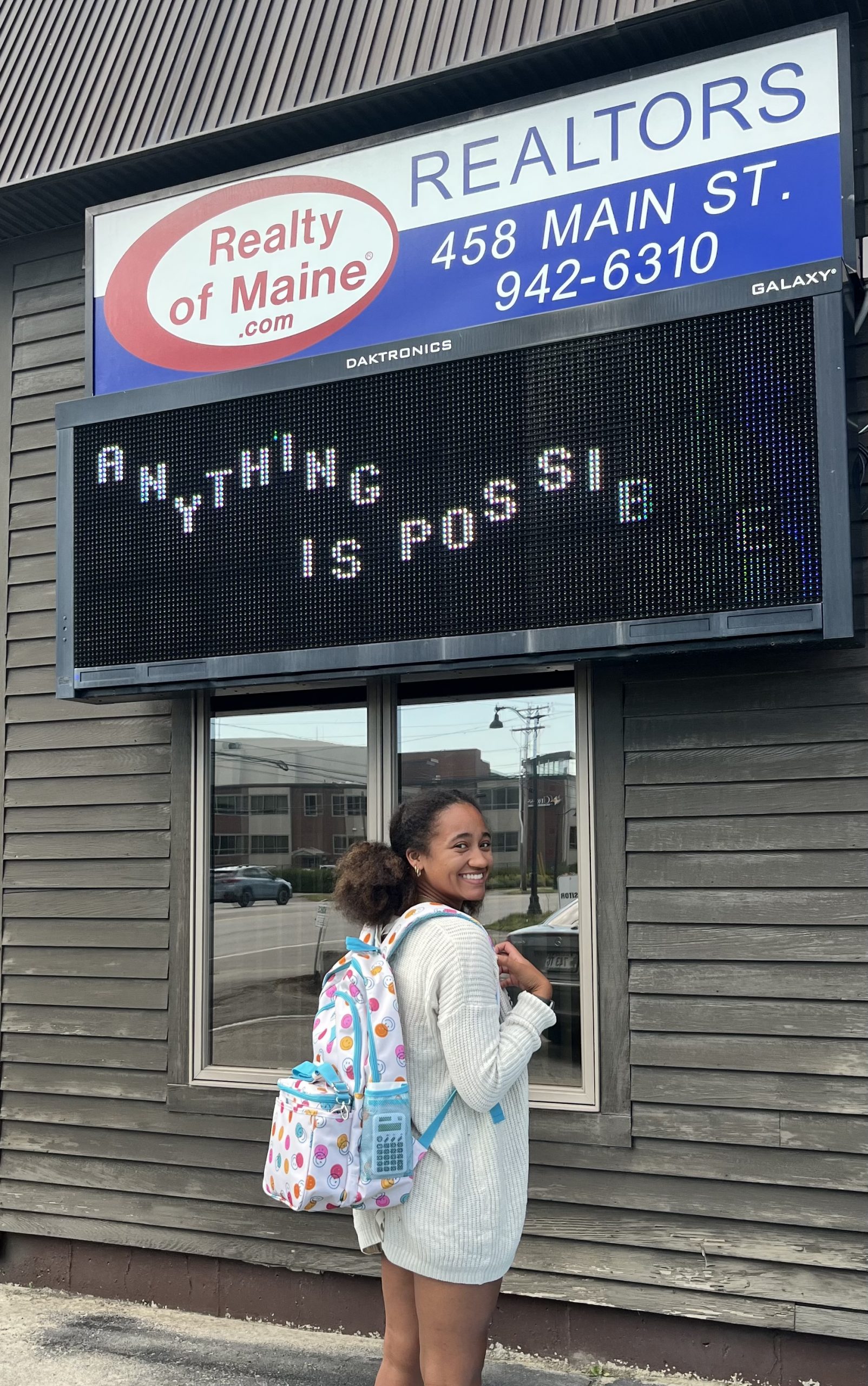 Malaya standing in front of Realty of Maine with a colorful backpack on.